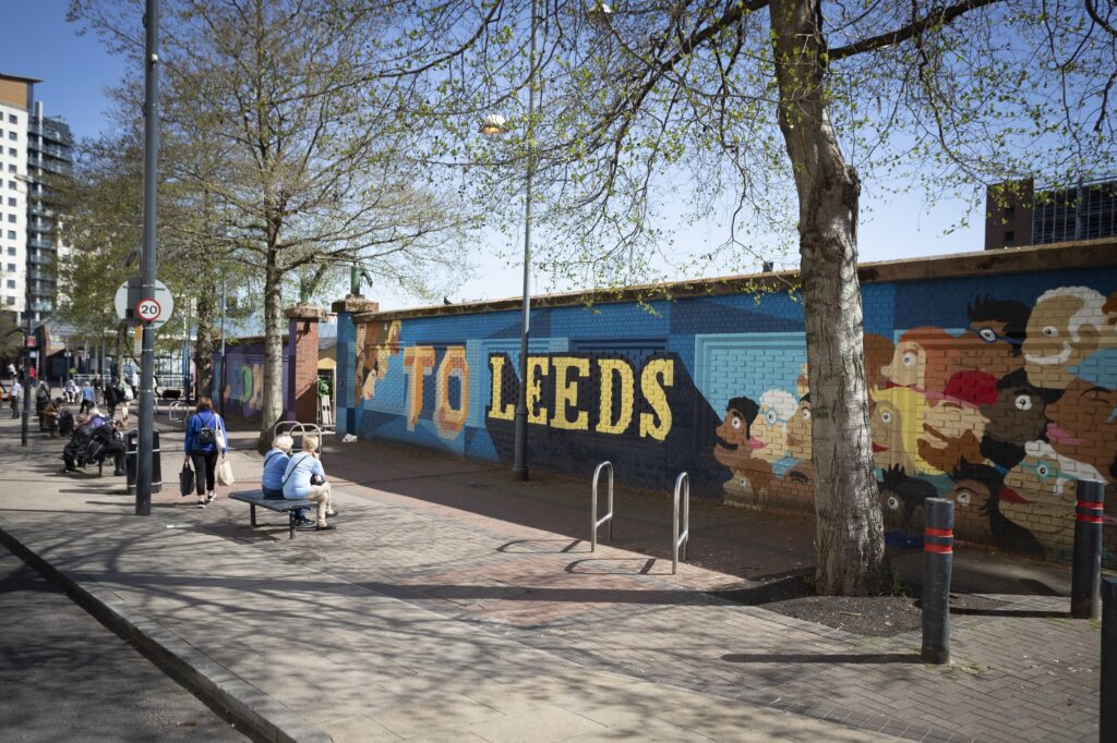a mural on a wall reading hell and welcome to leeds with peoples faces drawn, and people walking past