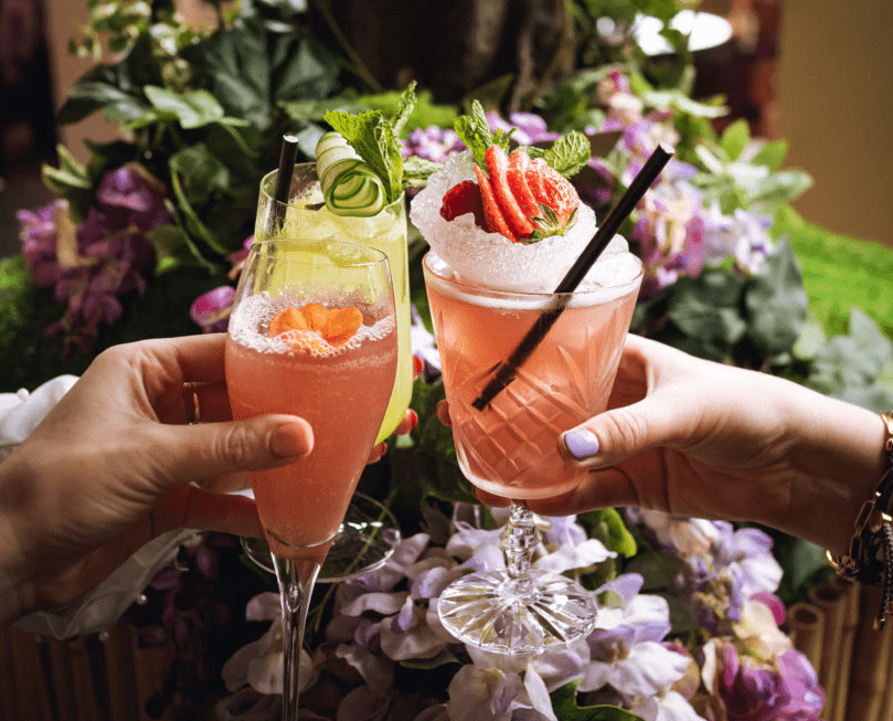 Two glasses of pink cocktails held up infront of purple flowers.
