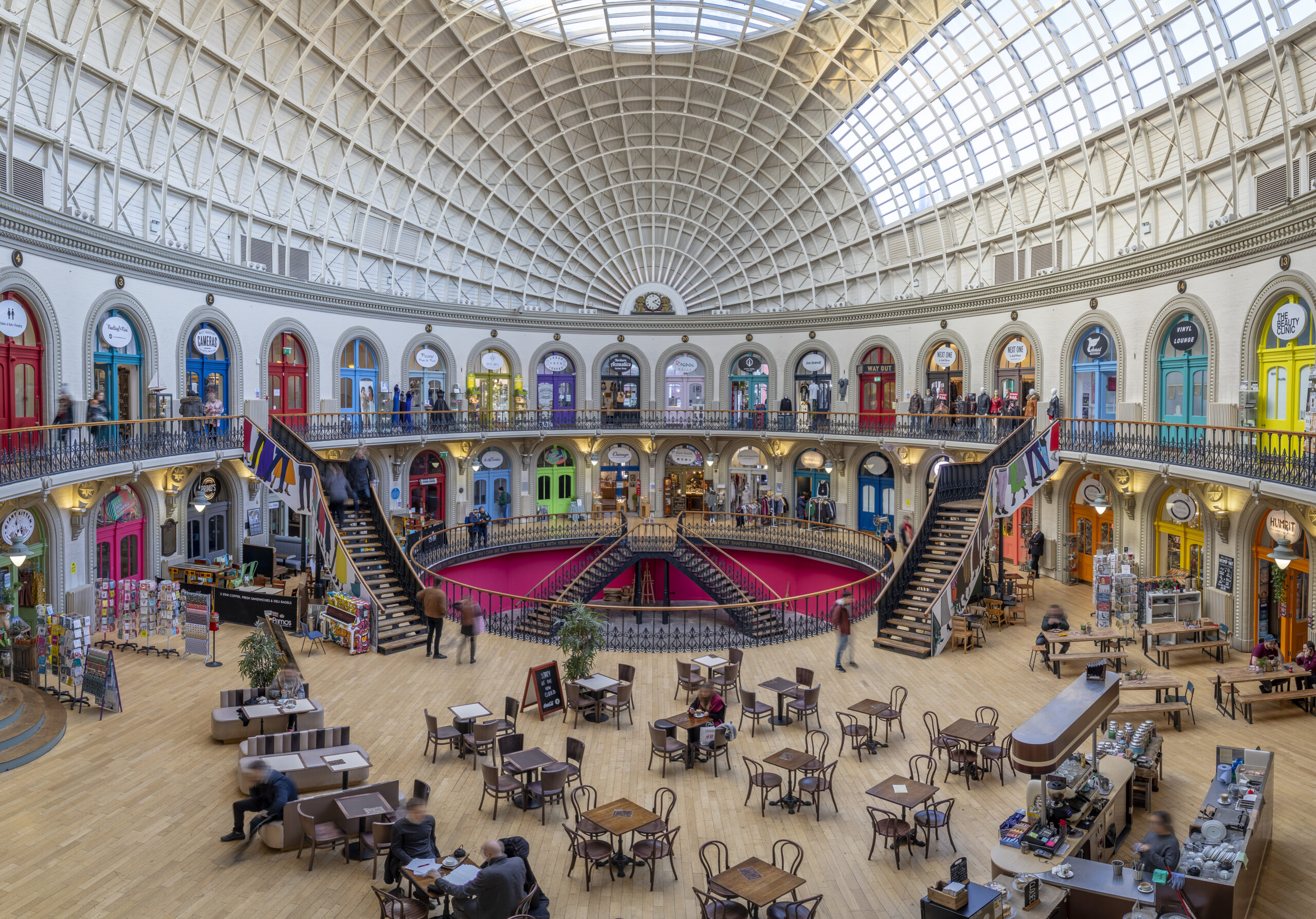 An interior view of the iconic Leeds Corn Exchange - a domed roof, colourful shop front entrances and staircases.