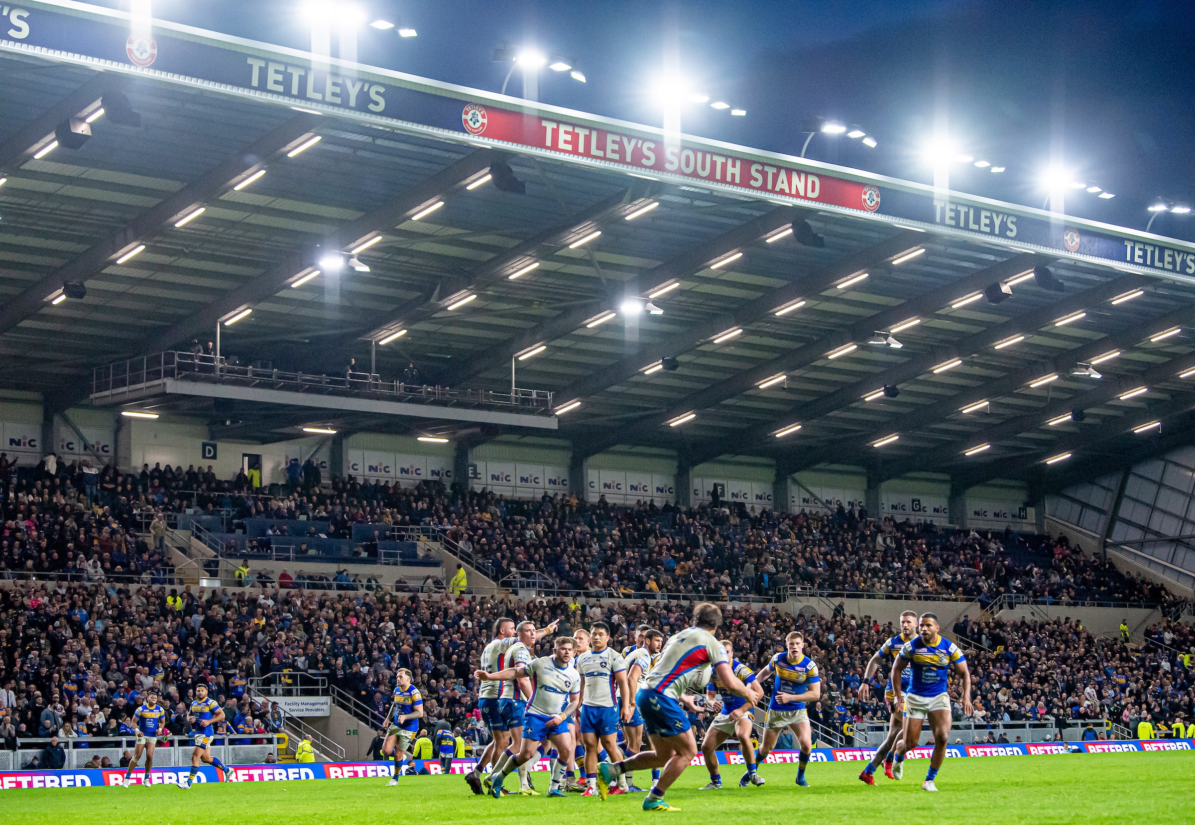 a rugby game in a stadium at night with large flood lights and players on the field