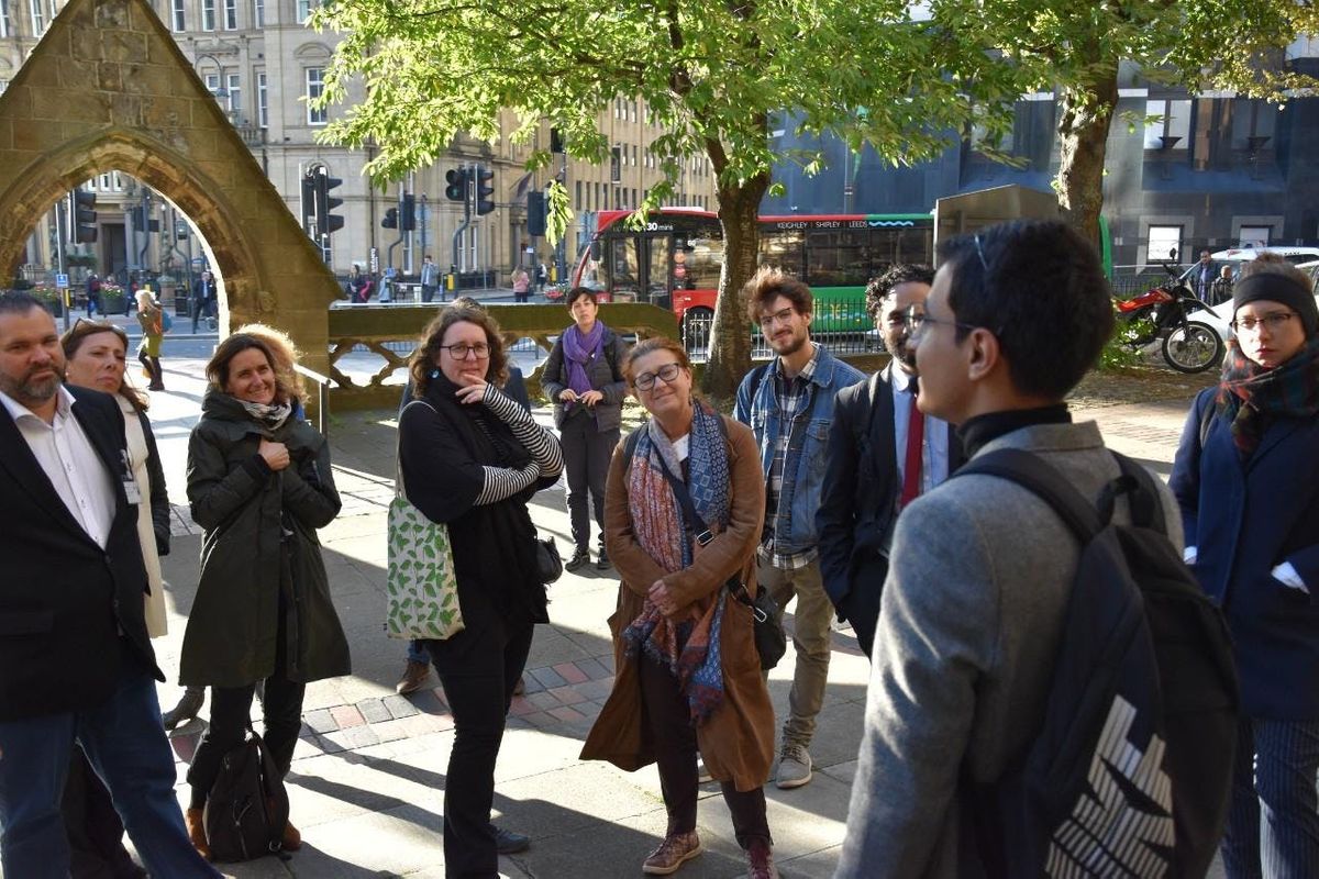 A group of people stand outside an entrance way listening to the tour leader talking, it is sunny.