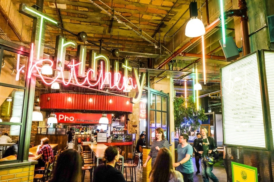 people walking inside a shopping centre food hall with the words trinity kitchen in neon lights above them