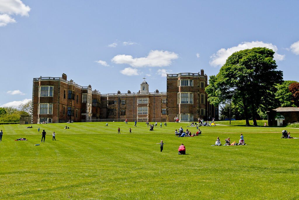 Exterior view of Temple Newsam, with lawn in front on a sunny day. People are sitting in groups enjoying the weather.