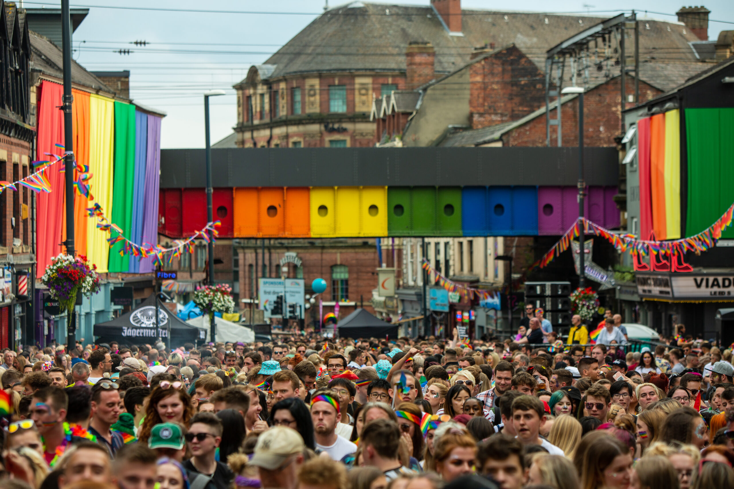 Photograph of crowd celebrating at Leeds Pride parade, beneath bridge painted in rainbow colours on Briggate.