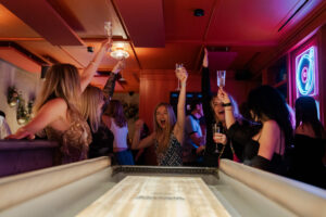 people cheering at the end of a shuffleboard wooden table holding up glasses