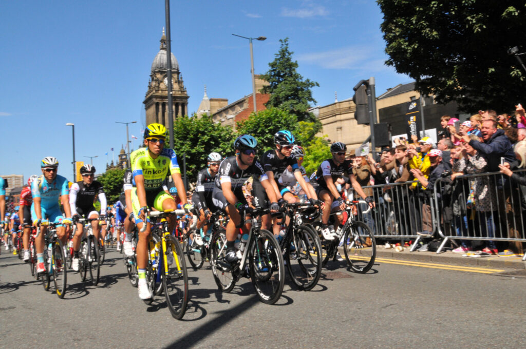 cyclists in professional gear in front of the Leeds Town Hall, a large victorian building, with people cheering them on
