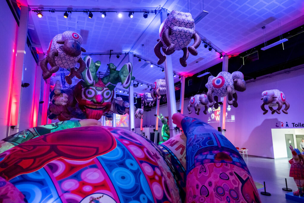 A vibrant indoor art installation featuring large, whimsical inflatable creatures with colorful patterns and abstract shapes, suspended from the ceiling.