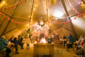 inside a tent with people stood around a fire and christmas lights