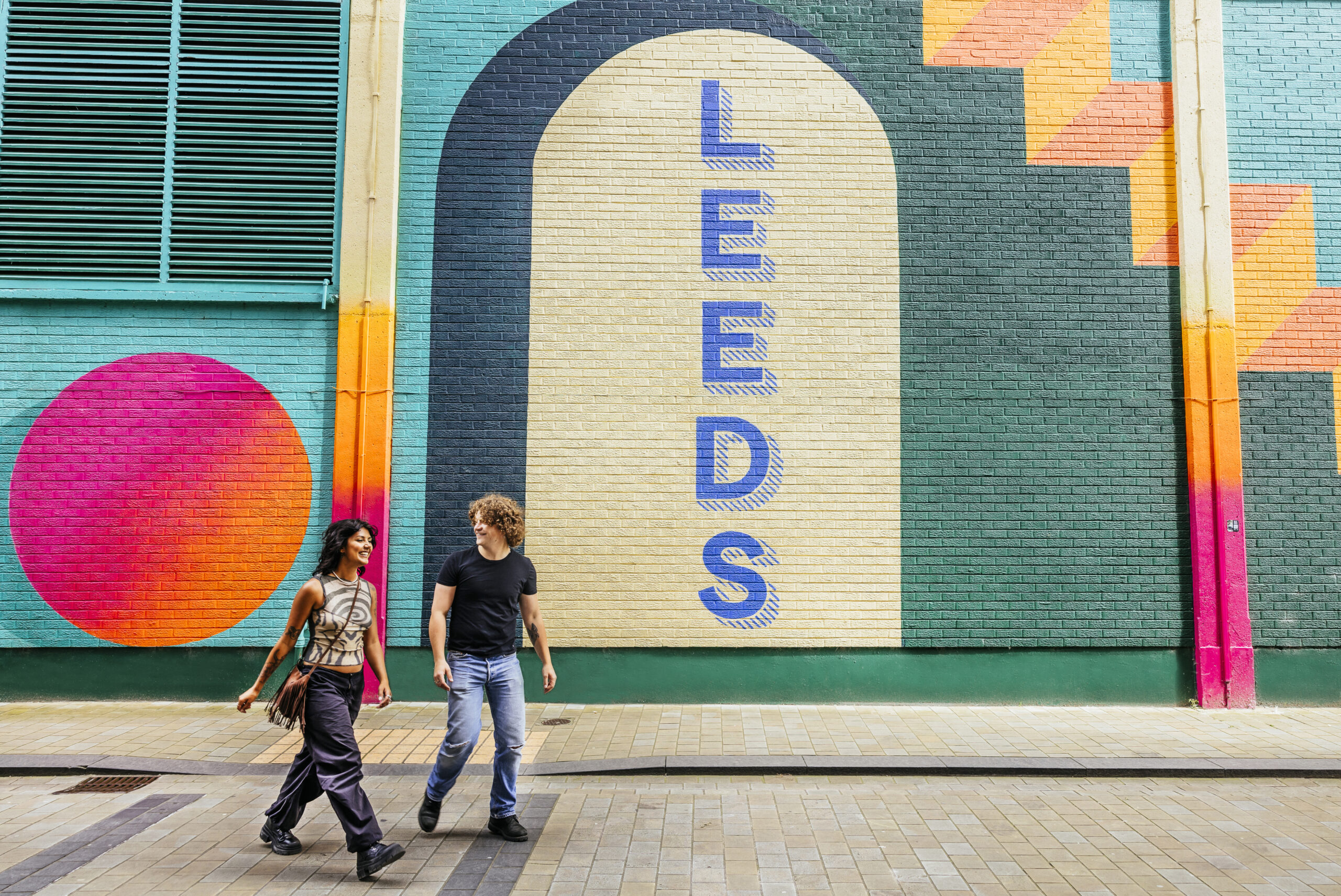 A young man and woman are smiling at each other as they walk past a brightly coloured mural with letters that spell out Leeds.