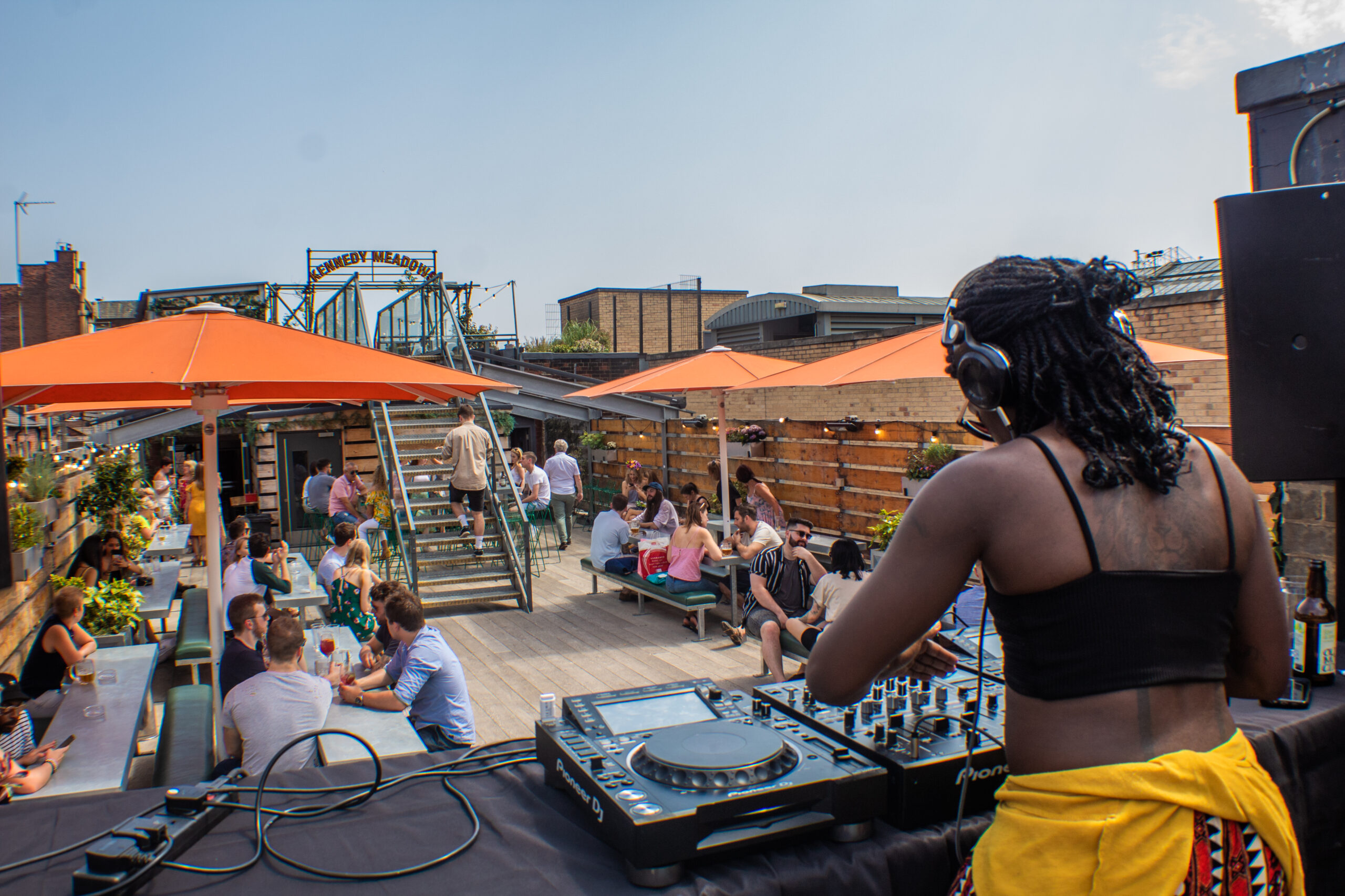 Groups of people enjoying a woman who is DJ's on the rooftop of Headrow House in the sunshine.
