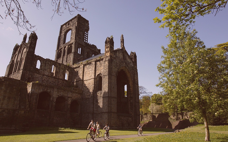 Image of the ruins of kirkstall abbey on a sunny day with blue skies, surrounded by grass and trees with two cyclists in the forefront.