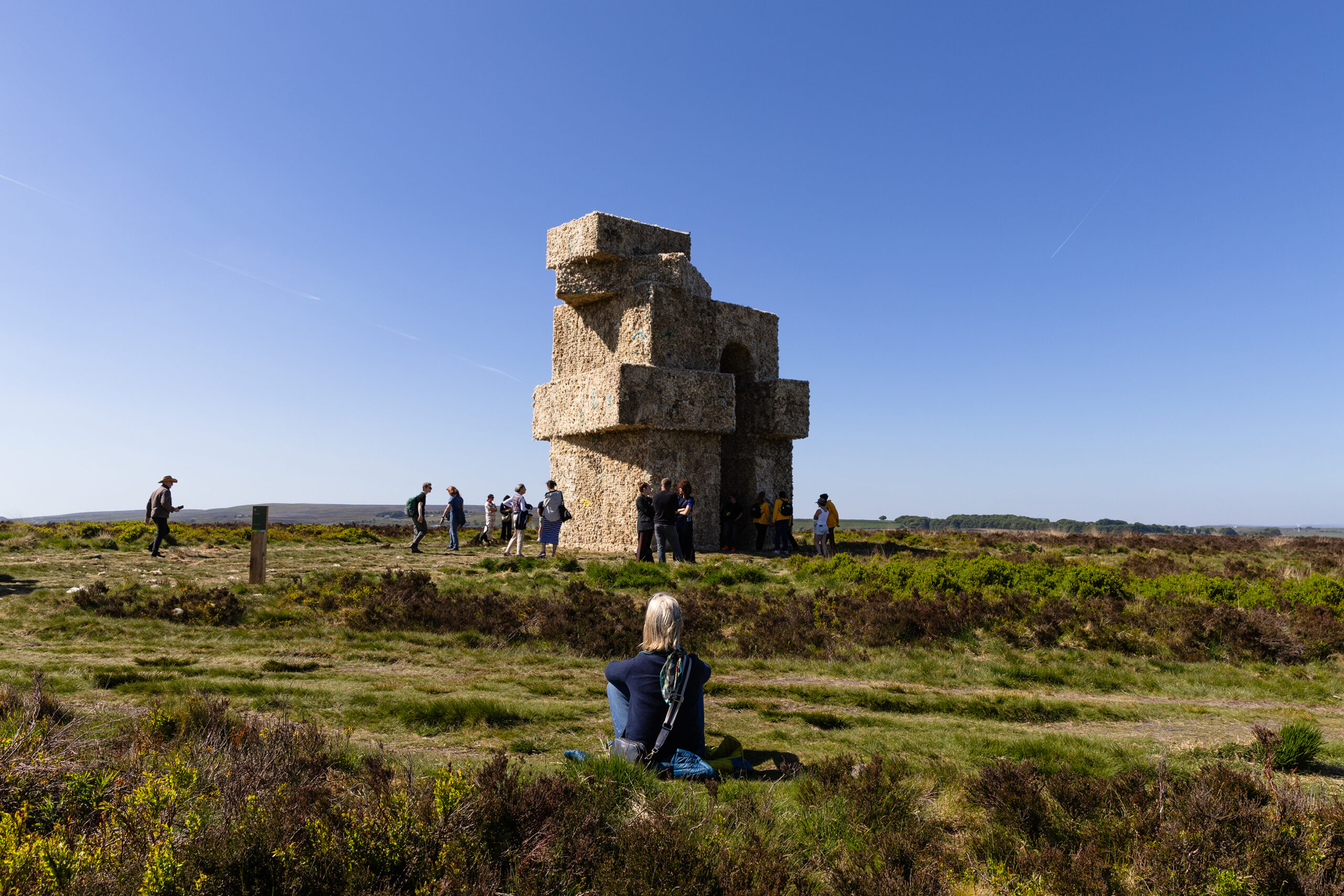 Photograph of a modernist concrete stacked sculpture of cubes surrounded by grass, in the foreground a girl sits and looks at it with her back to the camera.