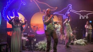 group of people on a stage singing and playing instruments with dinosaur lights