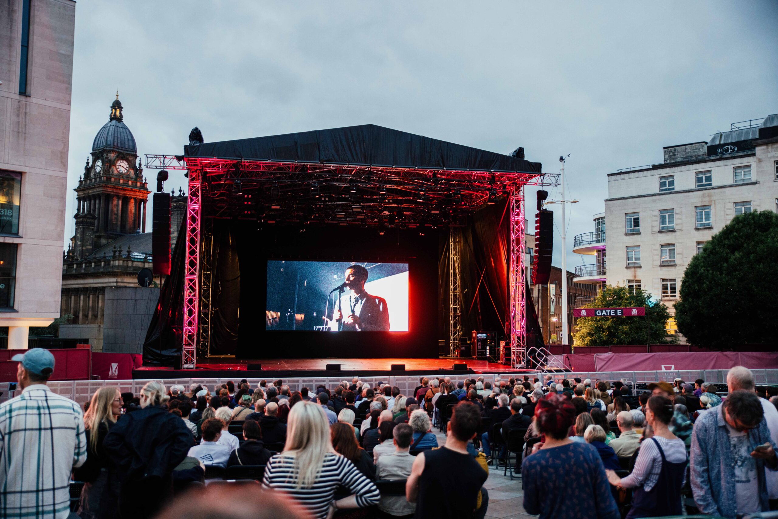 large group of people sat watching a movie on an outdoor screen with buildings in the background