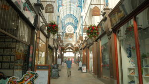 a victorian arcade with blue arches, flower pots hanging and a person walking through