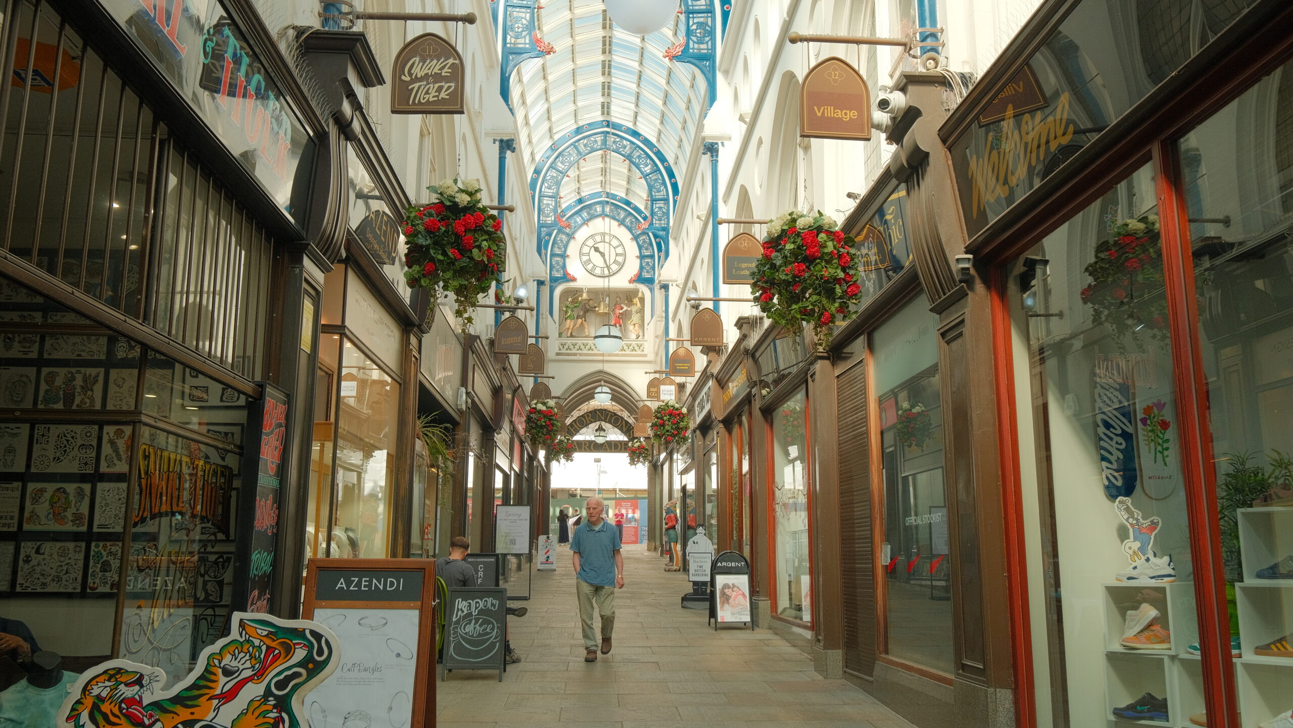 a victorian arcade with blue arches, flower pots hanging and a person walking through
