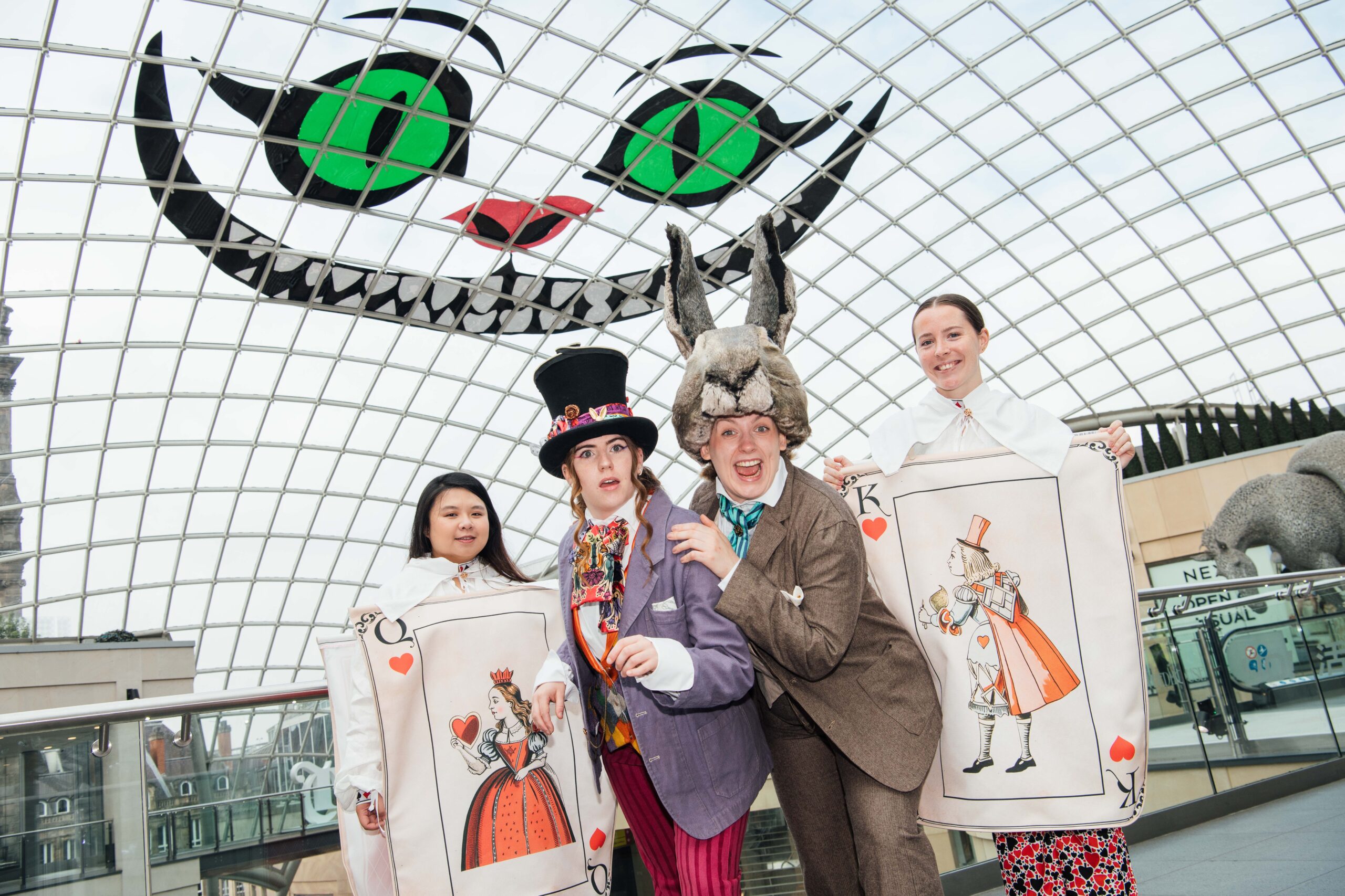 4 people dressed up as playing cards, a mad hatter and a rabbit in front of a ceiling with a large grinning face