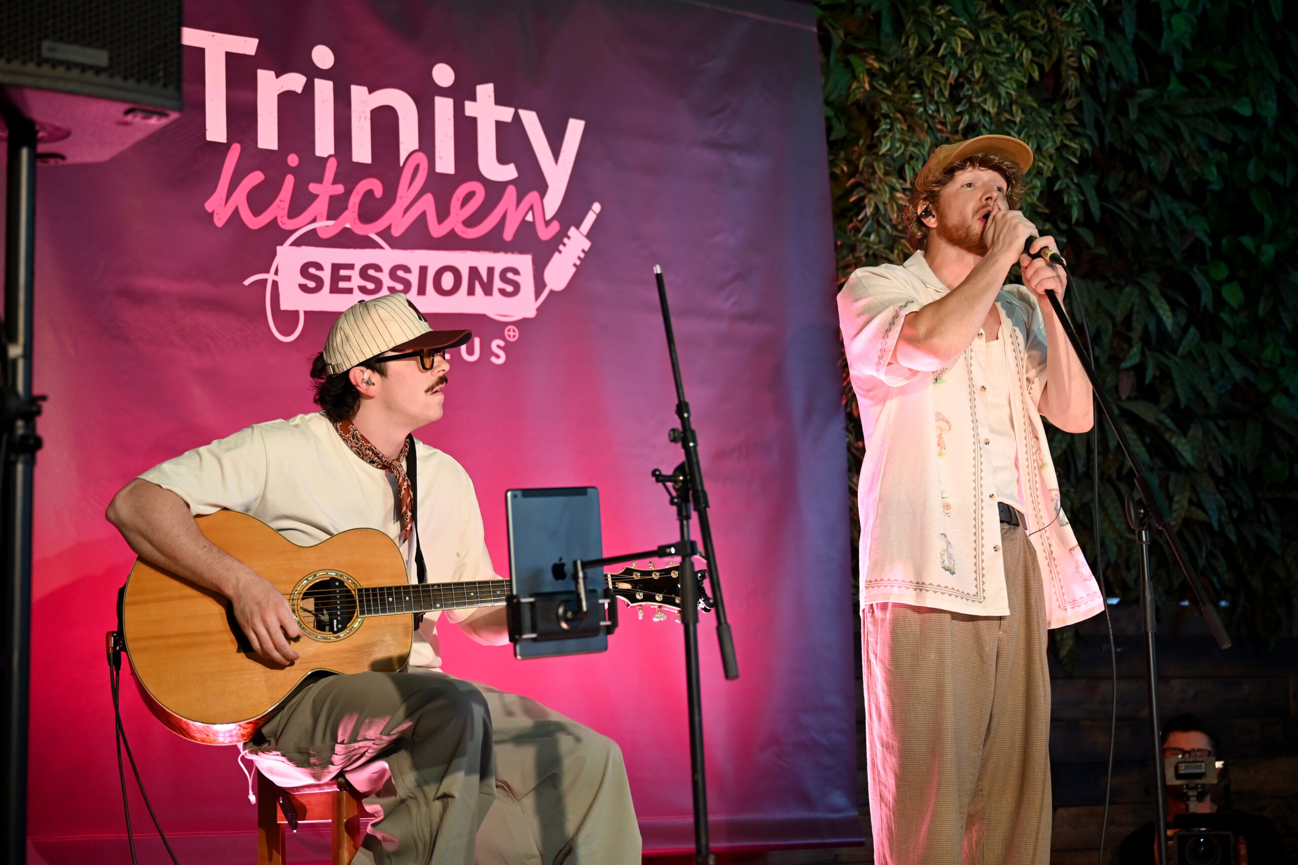 two people on a stage, one stood up singing and one sat playing an acoustic guitar with a sign behind saying trinity kitchen thursday sessions