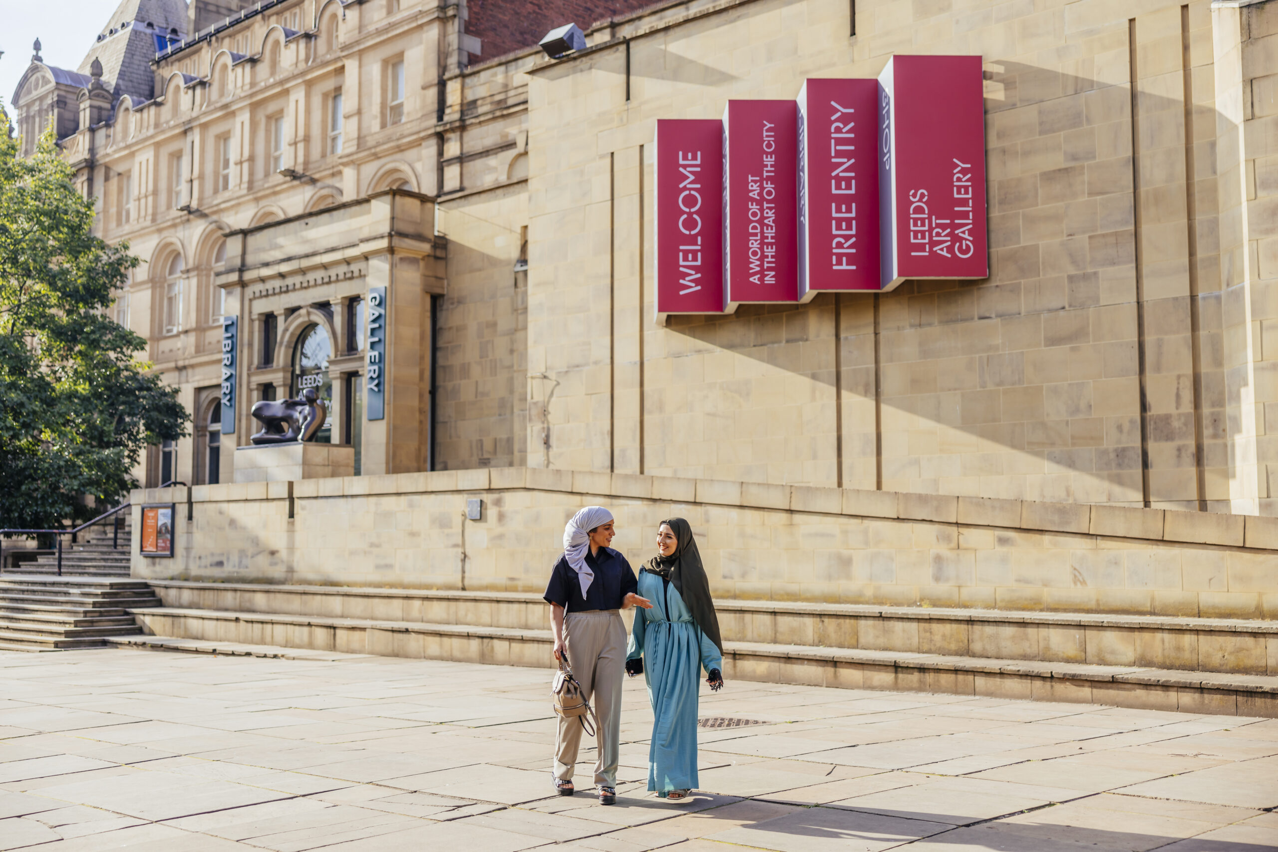 Two women arm in arm walk past Leeds Art Gallery in the sunshine.