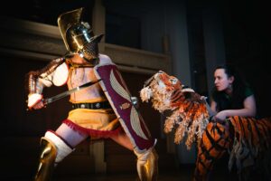 an actor in an ancient roman gladiator outfit, with a helmet, sword, shield, leg greaves, fighting a person holding a tiger puppet