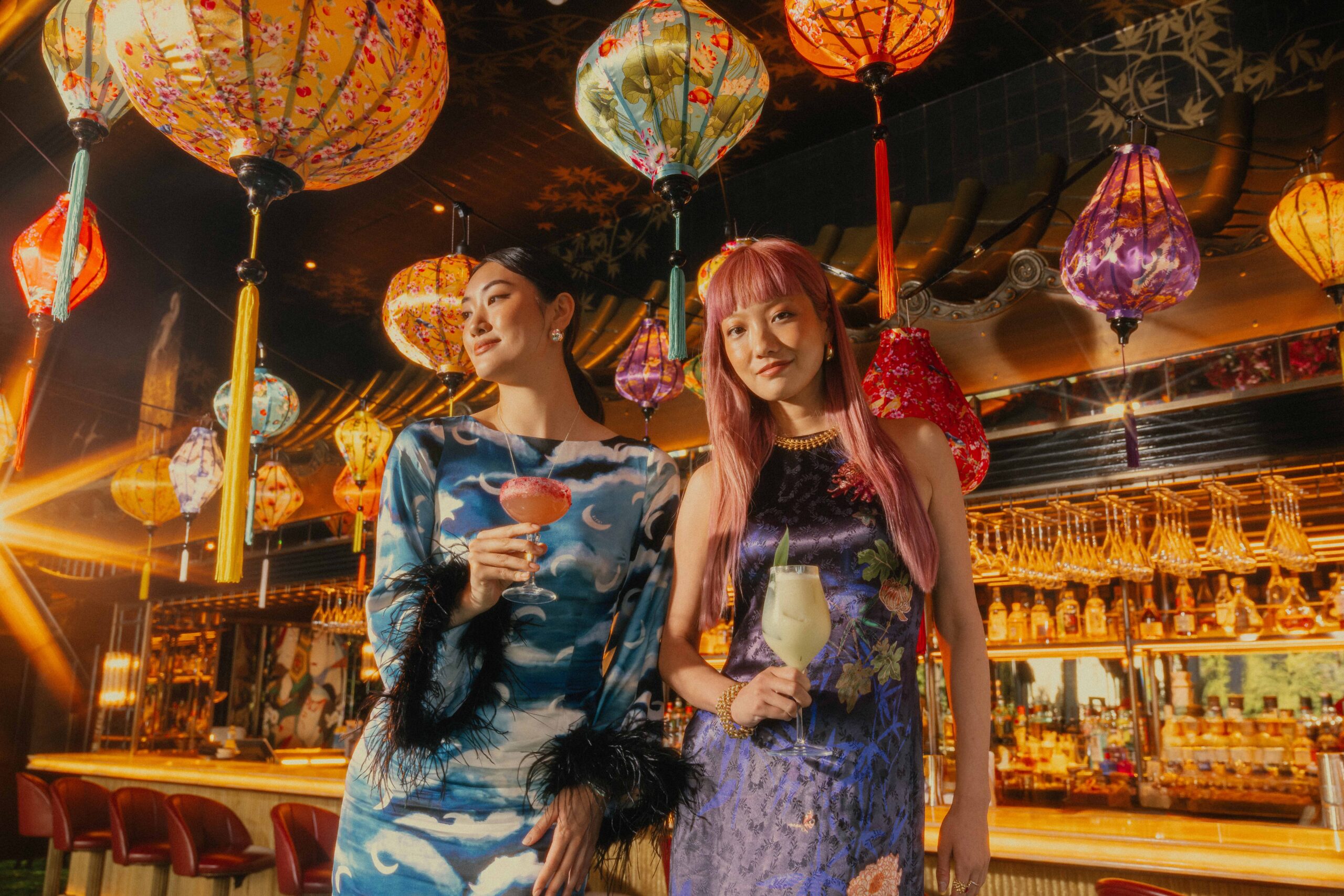 two women in silk dresses holding cocktails in a room decorated with ornate paper lanterns