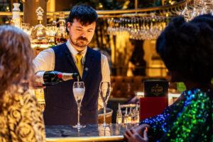 a man in a waistcoat pouring champagne for two ladies at a decorated bar