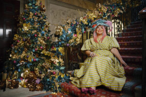 a christmas tree at the bottom of a decorated staircase and a woman dressed colourfully sat next to the tree