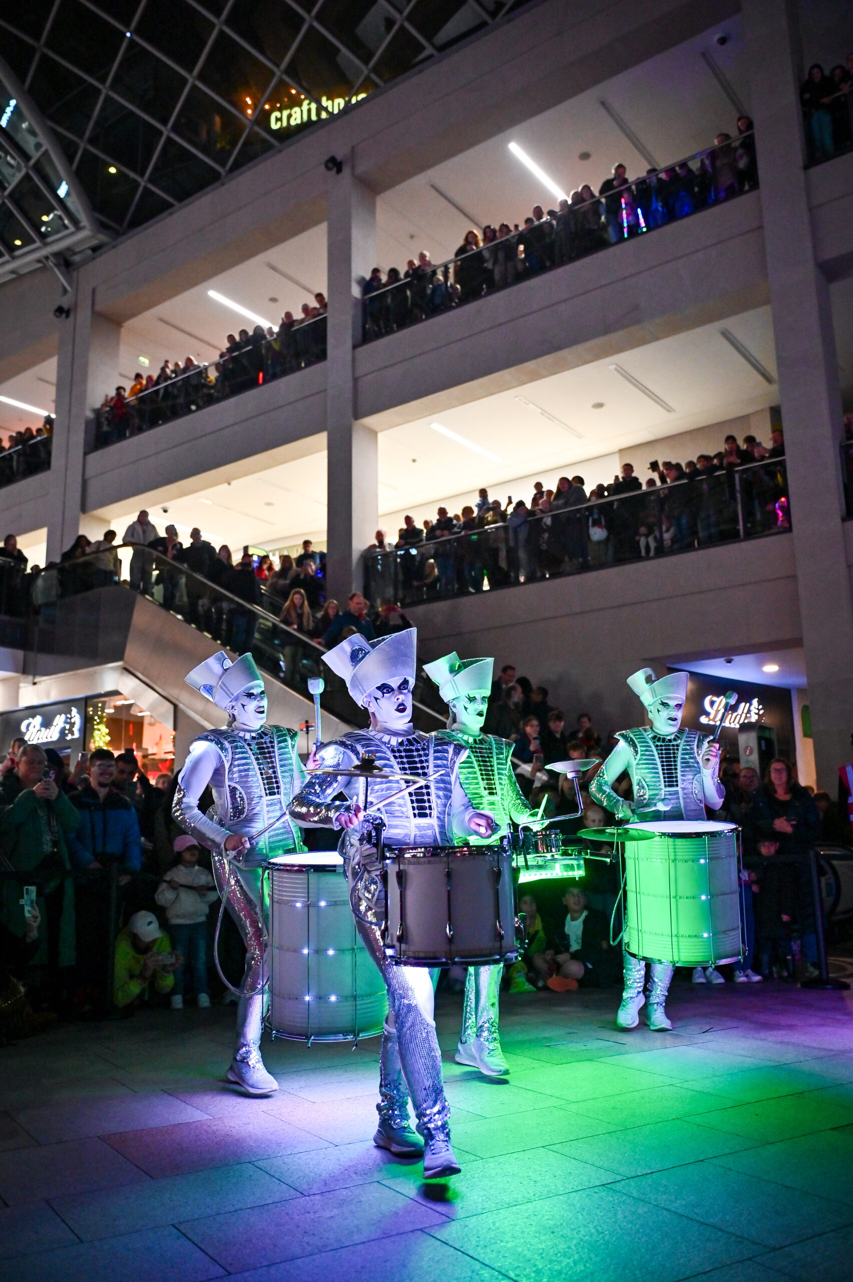 a group of people in silver costumes carrying drums all lit up in a dark shopping centre