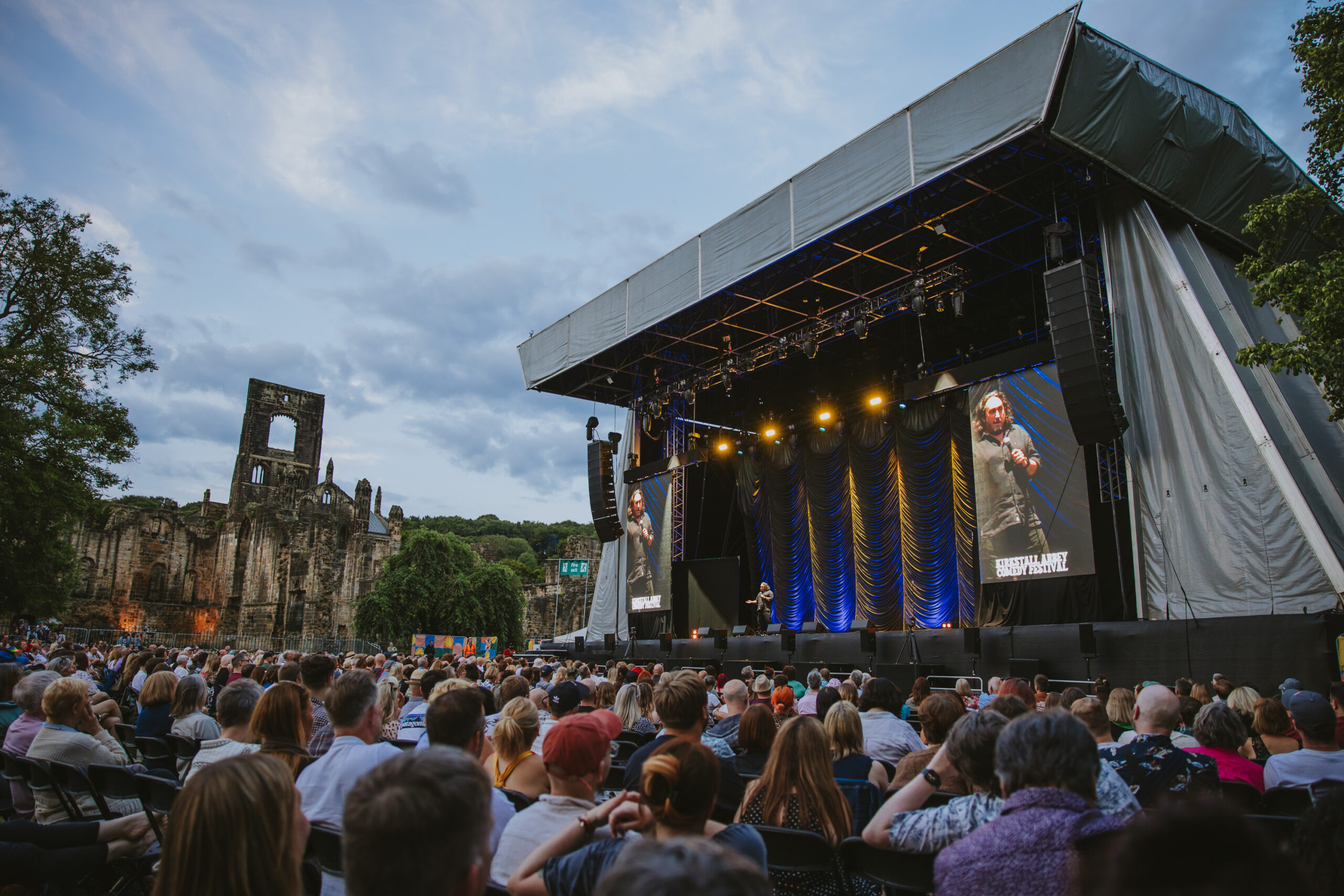 a large crowd of people watching performers on a stage with abbey ruins in the background