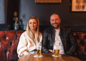 a couple sat a table with pints of beer in front of them on a wooden table 