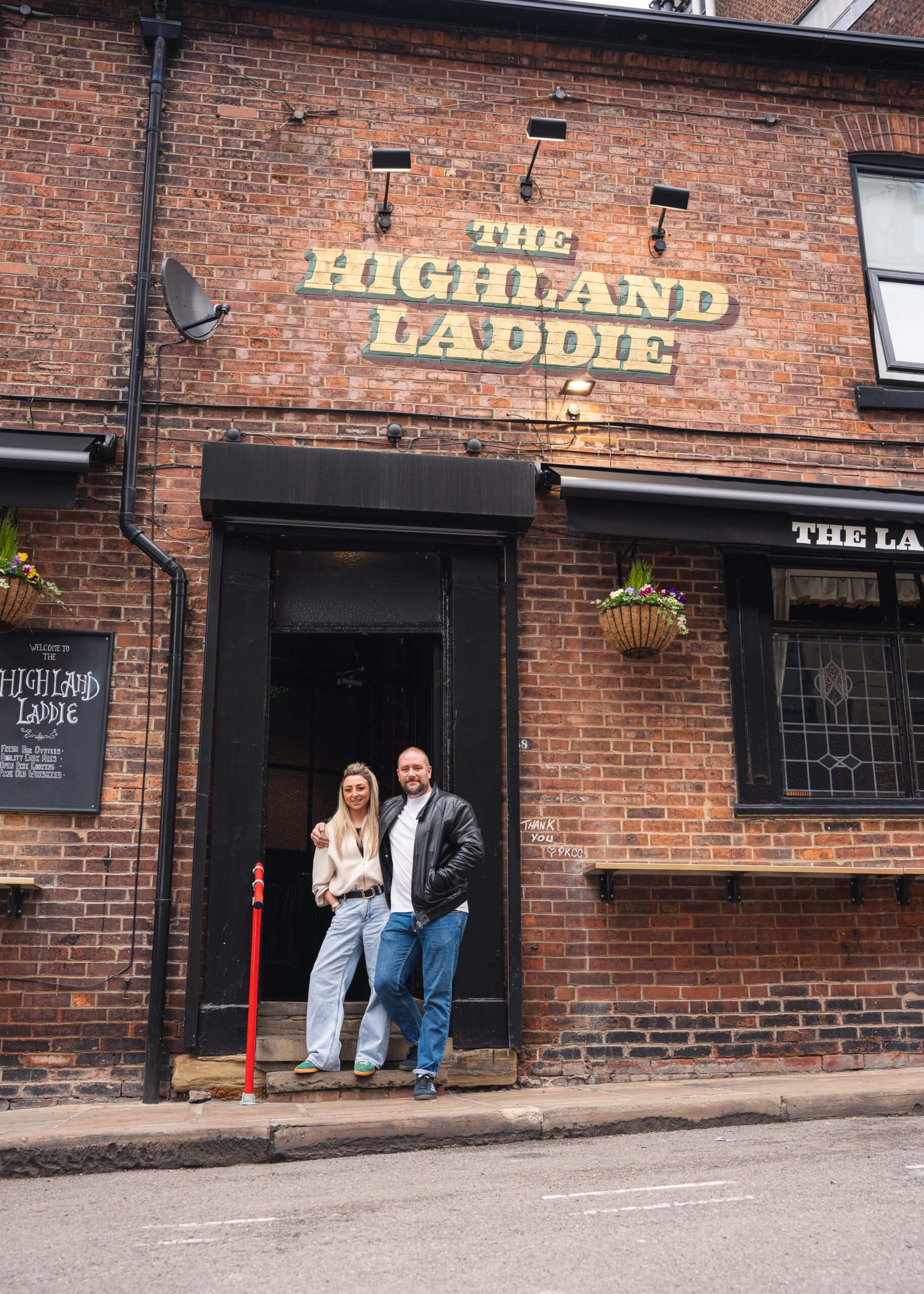 two people stood on the doorstep of a brick building with the words the highland laddie above them