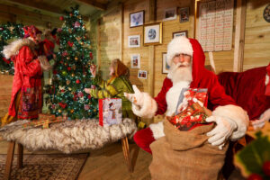 santa claus sat in a cosy wooden cabin with two elves behind decorating a christmas tree