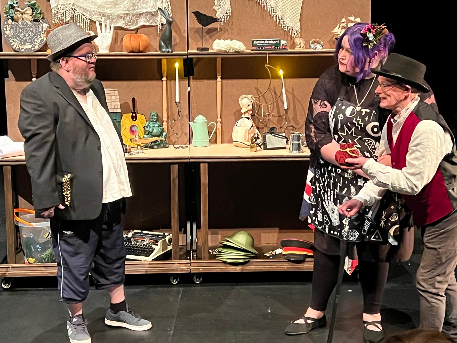 a couple of people stood in a rehearsal space with shelves full of random items behind them