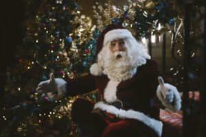 father christmas with a big white beard and red and white outfit in front of a traditional green christmas tree