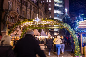 a crowd of people walking under a green archway with lights on it and a star above, a christmas tree in the background