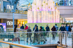 a crowd of people in a large open shopping centre with a modern christmas tree behind them with stars and tubes of fairy lights through them
