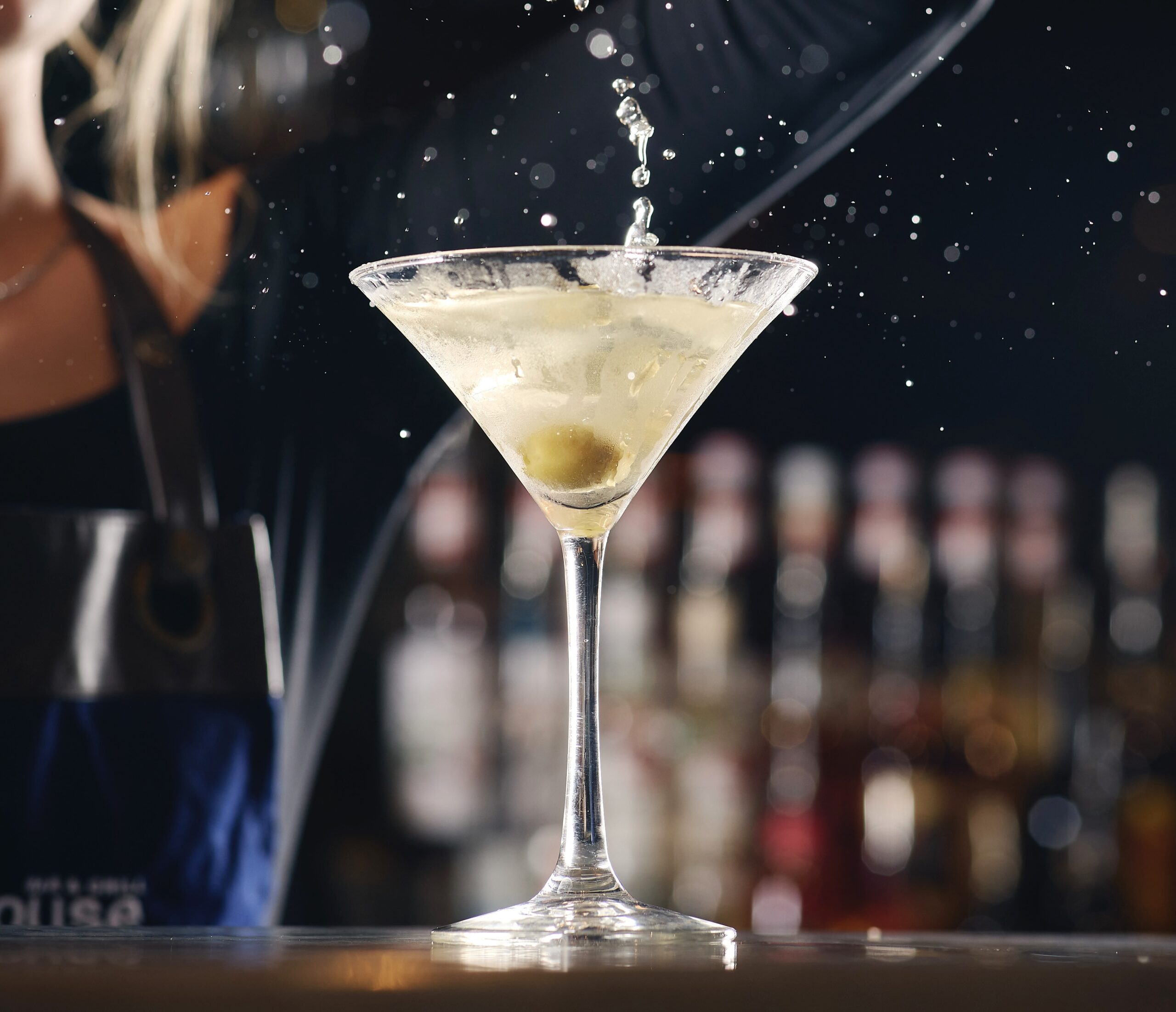 a martini glass with an olive in it and a bartender pouring into it