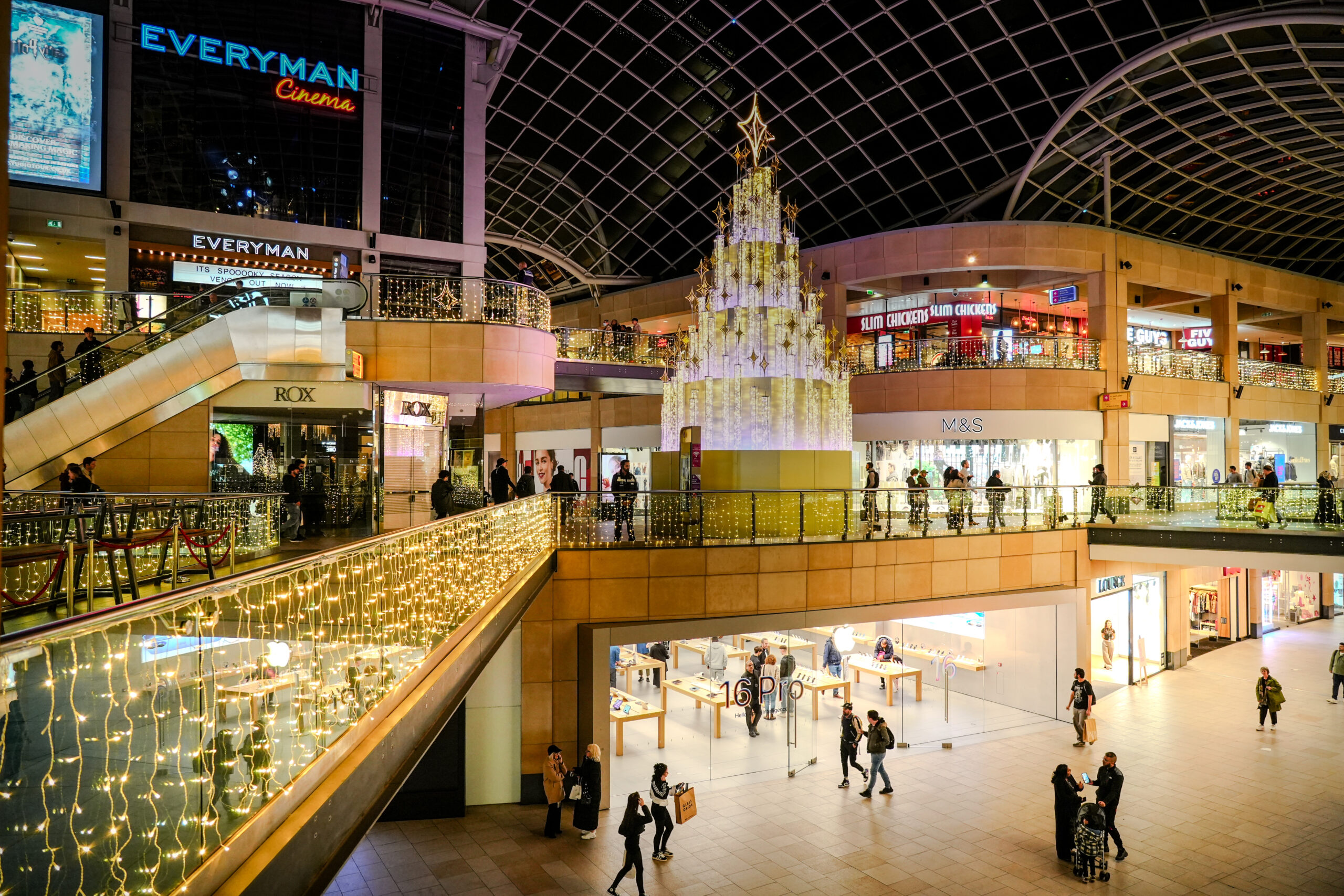 a large shopping mall with two floors, a glass roof, with a large sparkling christmas tree in the middle and fairy lights across the balconies