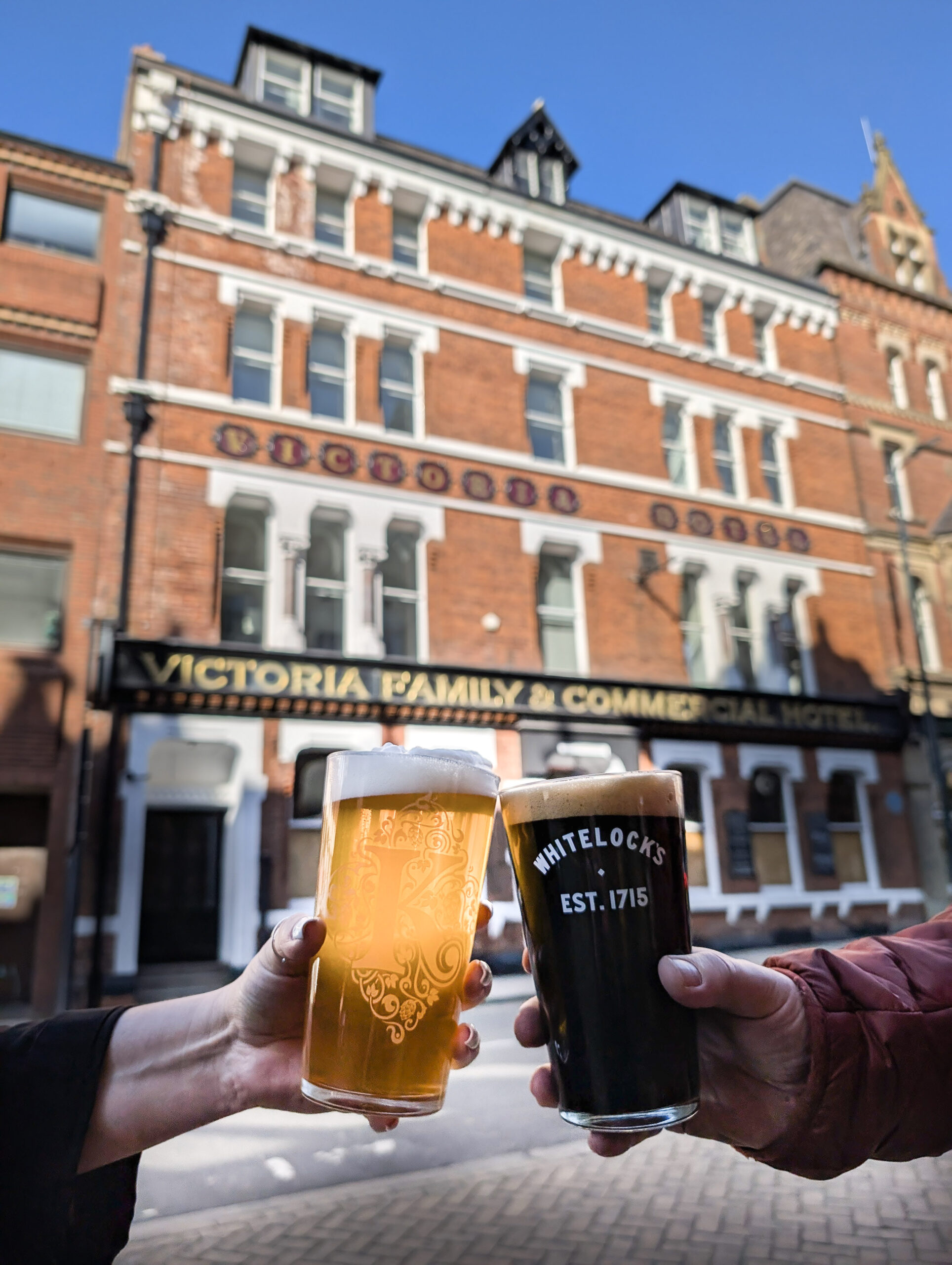 2 hands holding pints of beer in front of a building with an old fashioned sign saying victoria family and commercial hotel