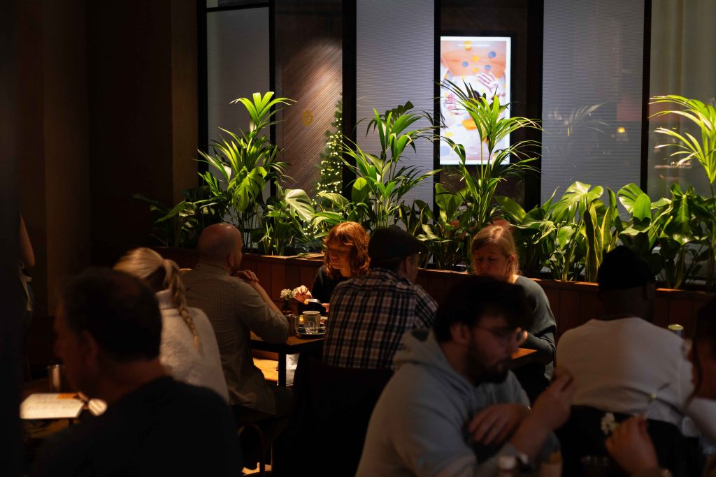 people sat in a cafe space with warm low lighting, green plants behind the seats