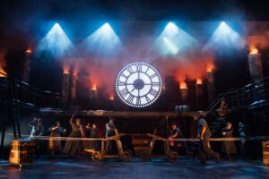 cast of actors on a large stage with a lit up analogue clock above them