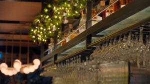glasses hanging above a bar with liquor bottles above and then green wreaths with fairy lights all under a warm light