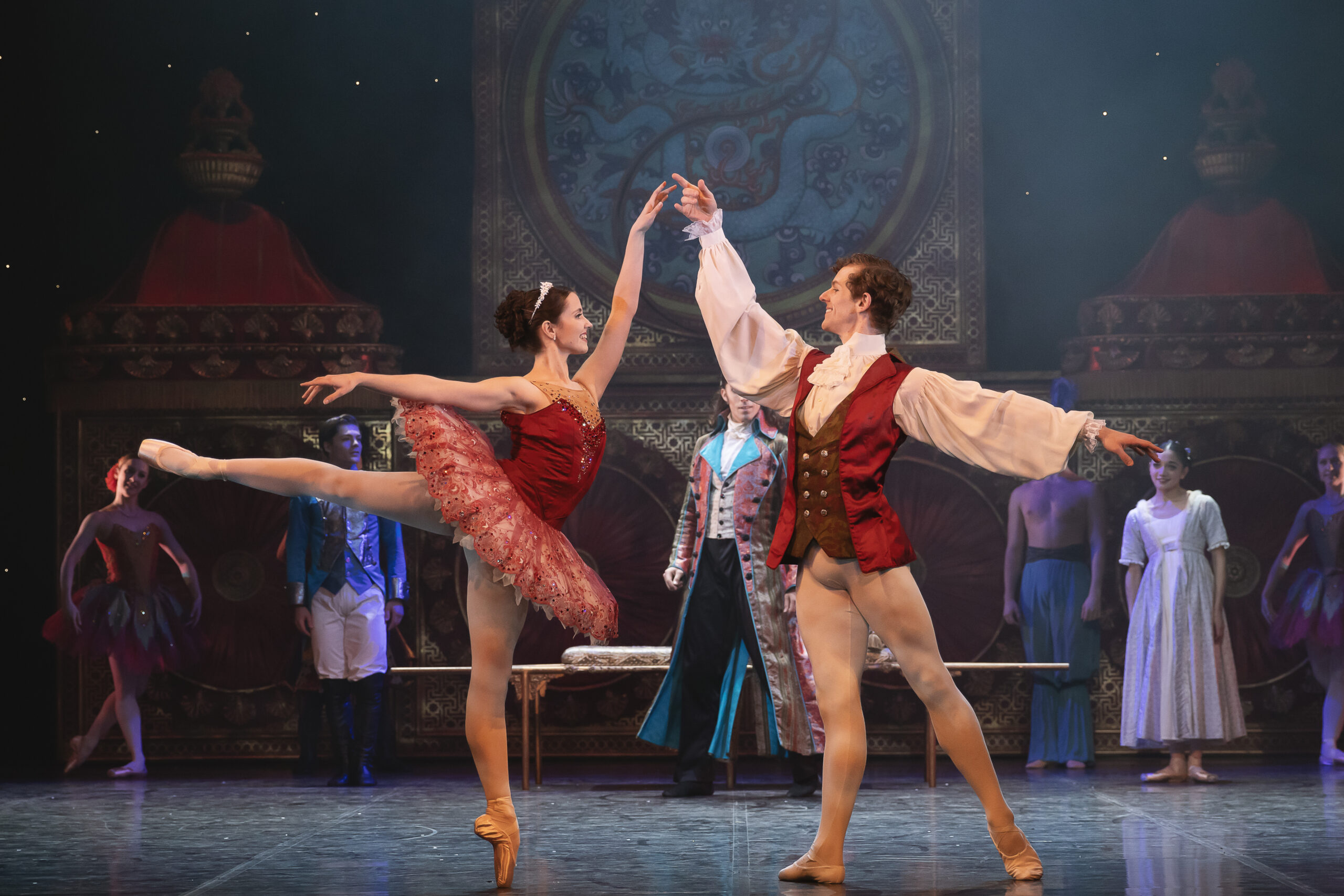 two ballet dancers in the foreground, one in a tutu en pointe, and another holding her hand whilst more cast watch from behind