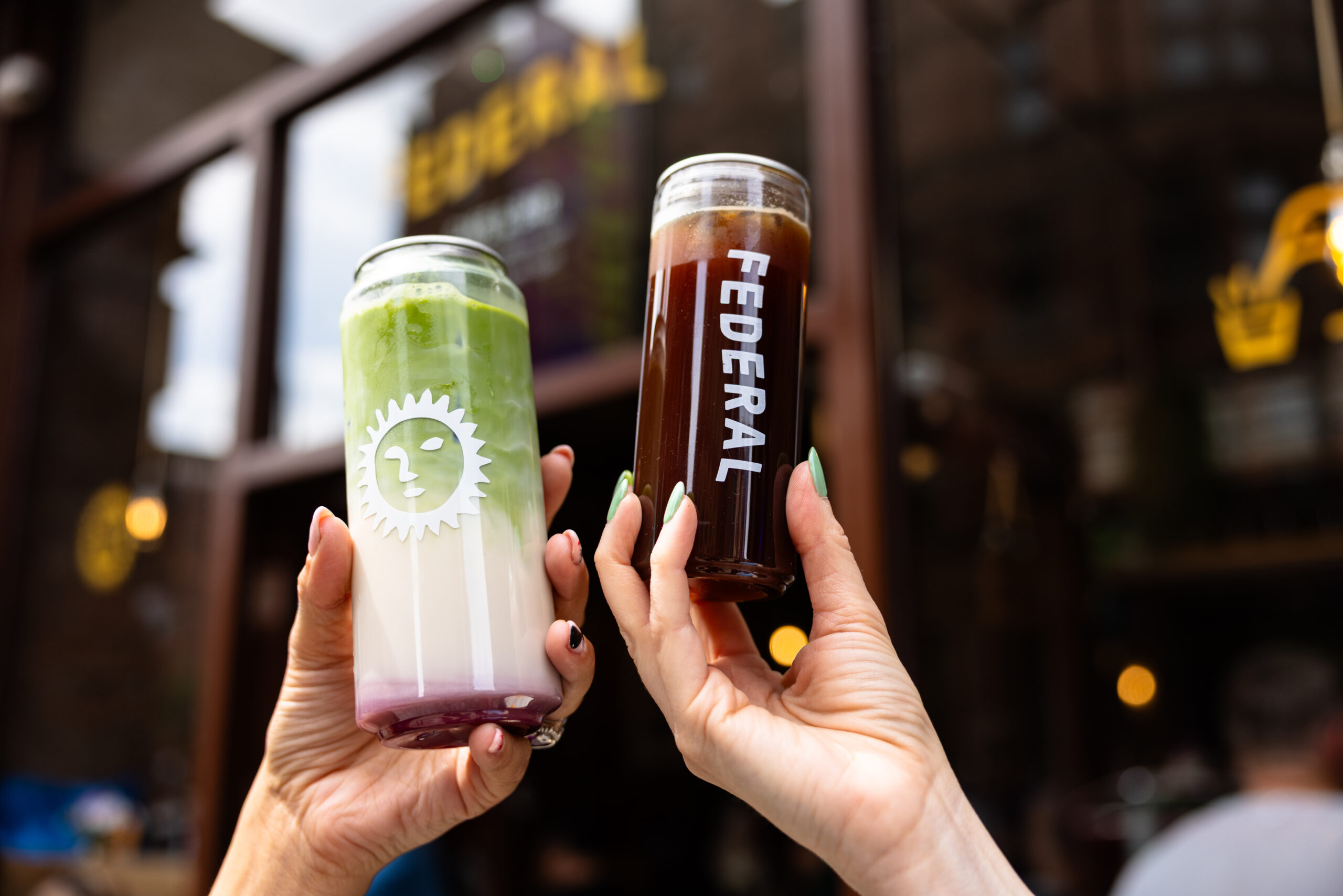 hands holding up two clear cans, one with a matcha green drink and the other a dark coffee, with the federal name on them