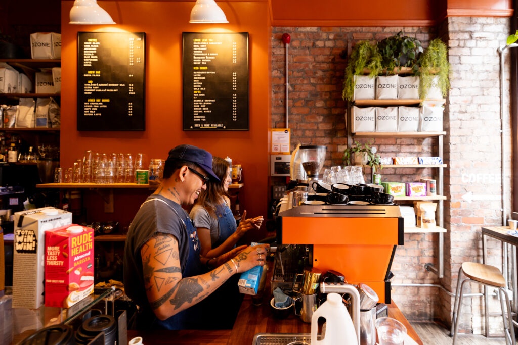 a cafe with orange walls, one exposed brick wall with green plants and merchandise on it, a couple of baristas making coffee at a large espresso machine