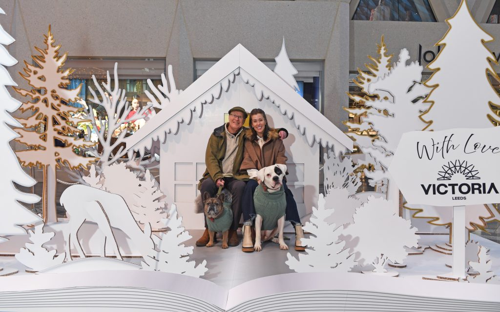 an installation which looks like a popup book of a snowy scene with trees, a cottage and deer, with a couple sat in the middle with two dogs