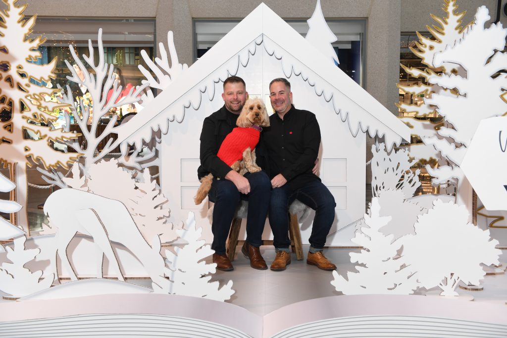an installation which looks like a popup book of a snowy scene with trees, a cottage and deer, with a couple sat in the middle with a dog on their laps