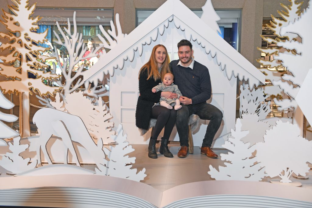 an installation which looks like a popup book of a snowy scene with trees, a cottage and deer, with a couple sat in the middle with a baby on their laps