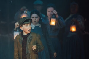 actors holding up old fashioned lanterns whilst a young boy stands in front singing