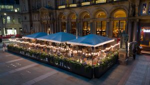 night time shot of a large building with a bar area out front under a tent with a large crowd of people inside and fairylights